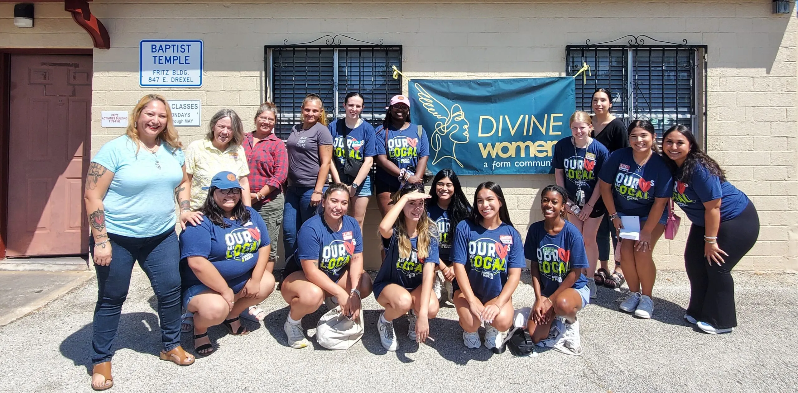 Divine Women members in matching shirts, smiling together outside in front of a Divine Women banner